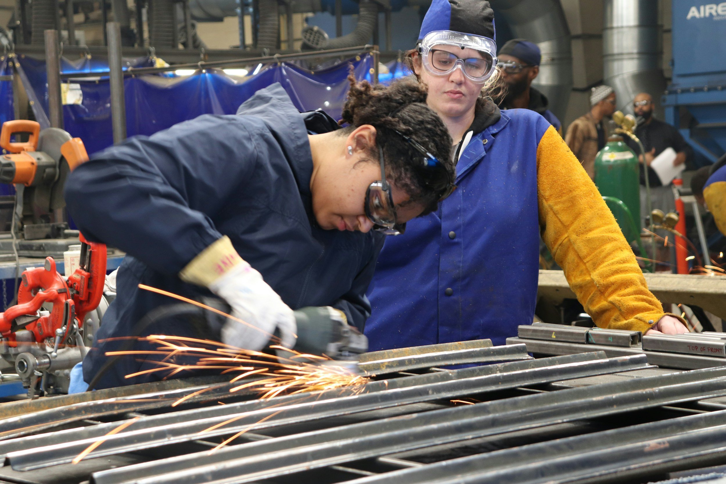 Zwei Frauen arbeiten in einer Werkstatt, eine schneidet Metall mit einem Winkelschleifer, während die andere zuschaut.